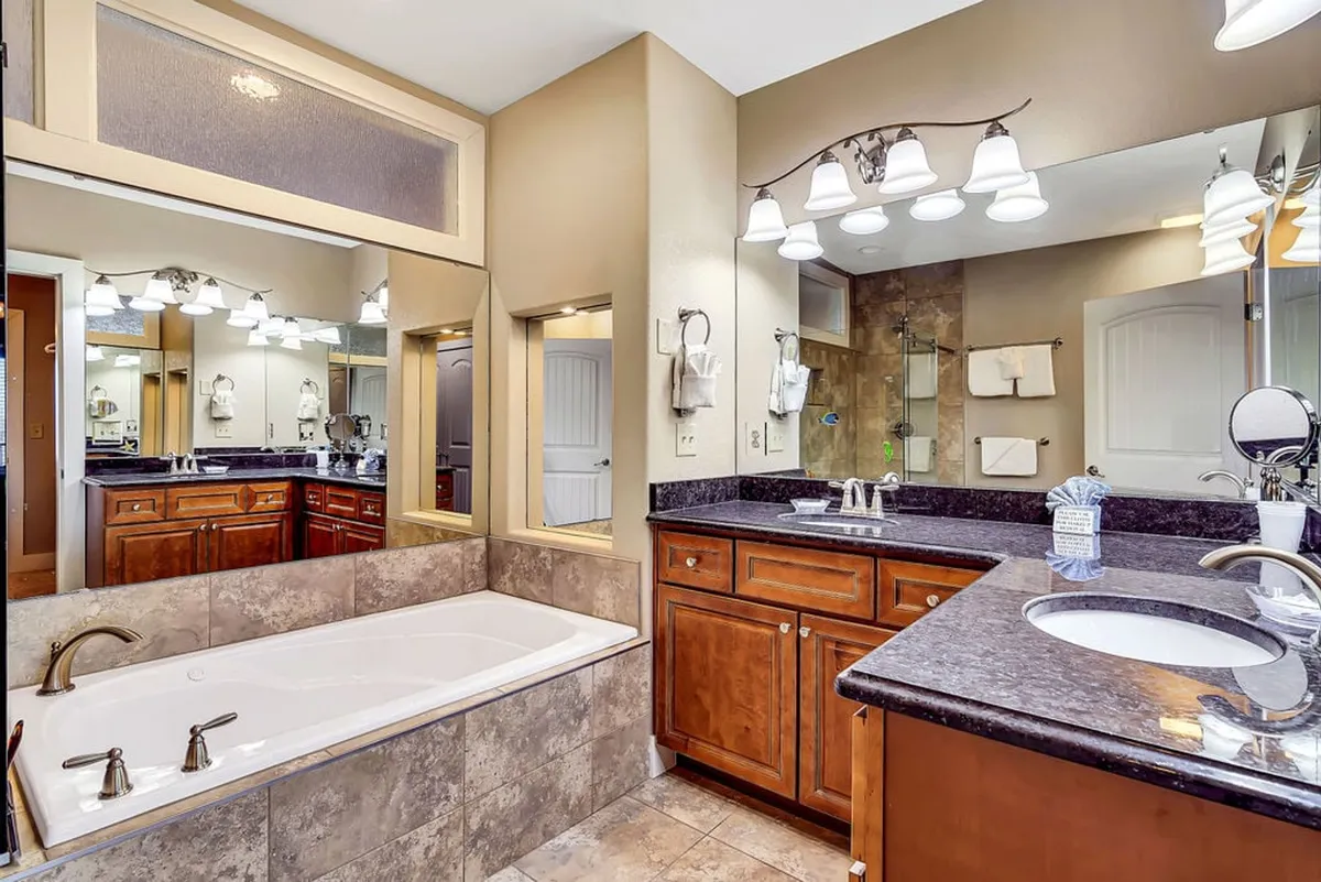 A luxurious bathroom scene with a deep soaking bathtub, double vanity sinks topped with large mirrors, and soft lighting fixtures. The space feels spa-like and inviting, ideal for self-care routines.