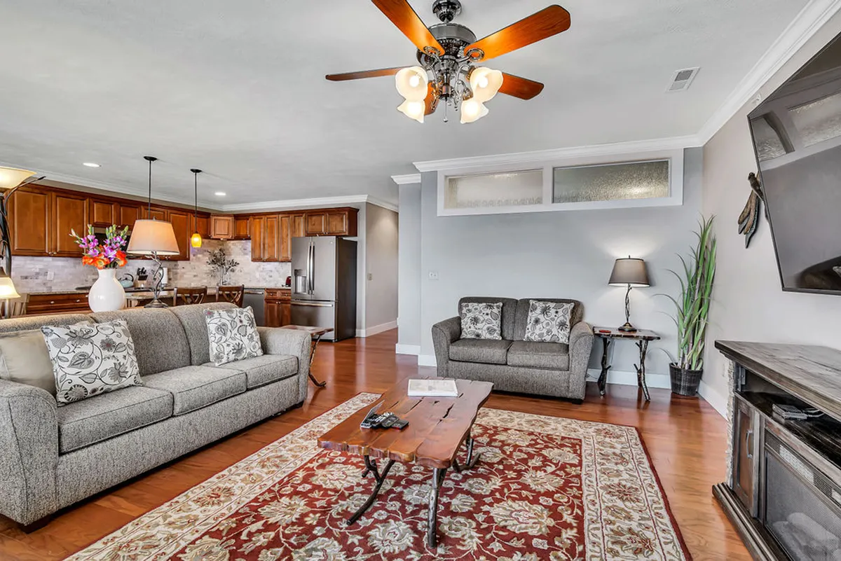 This image presents an open-concept living area that seamlessly connects the sofas, coffee table, chandelier, and kitchen area. The space is styled in a comfortable and inviting manner, with plenty of room for socializing, relaxing, and gathering with friends or family. The visual connection between the living and kitchen zones clearly supports a cohesive, communal atmosphere.