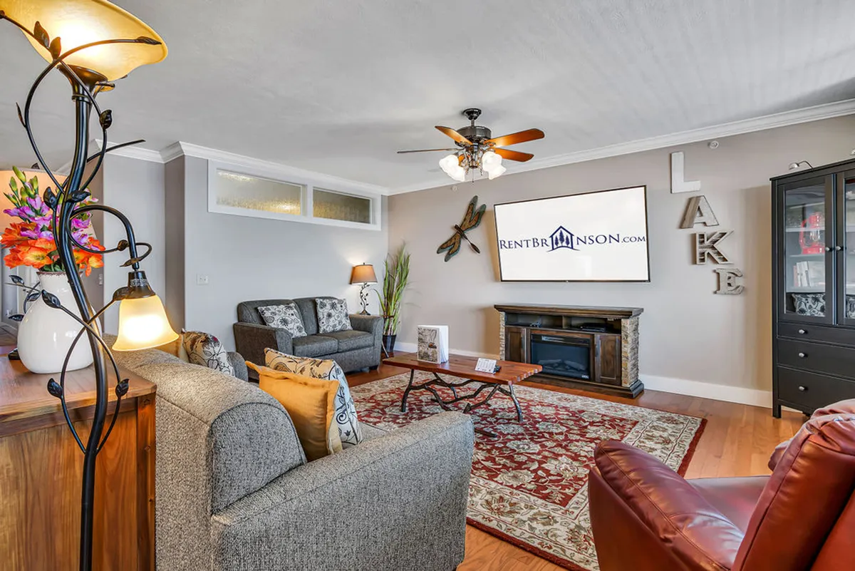 The image showcases a warm and welcoming living area featuring plush, comfortable sofas arranged around a sleek wooden coffee table and a large TV. Lake-themed wall decor adds a local touch, and the decorative rug and electric fireplace create a cozy atmosphere perfect for relaxing movie nights with family or friends.