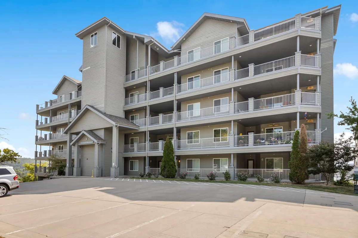 The building's entrance is step-free and inviting, with balconies adorned with plants and surrounded by lush greenery. The scene radiates a welcoming atmosphere and easy arrival, highlighting the seamless access to the home.